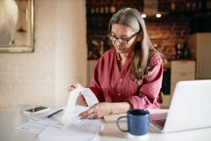 Serious focused middle aged female accountant in eyewear working from home providing outsource bookkeeping service, sitting at table with portable computer, calculating finances, doing paperwork